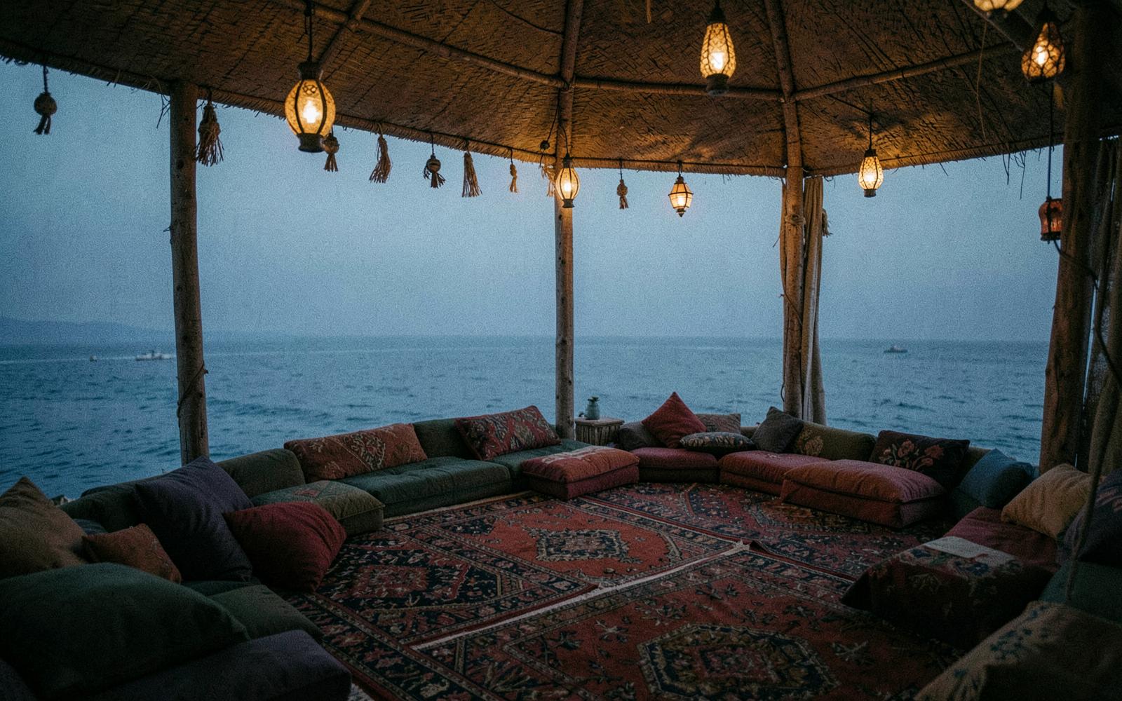 An outdoor majlis-style seating area with lanterns overlooking the sea at dusk